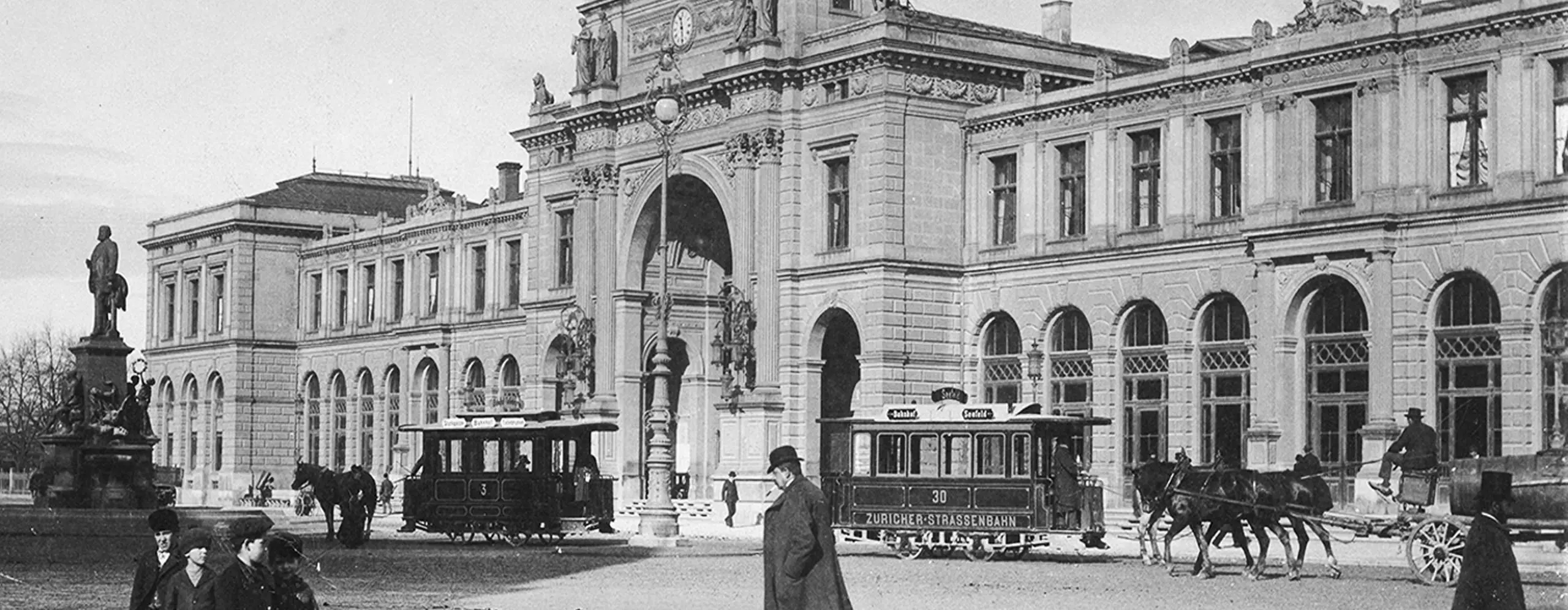 Historische Straßenszene mit Pferdebahn, mehreren Personen in zeitgenössischer Kleidung und einem großen Gebäude mit Bögen und Säulen.
