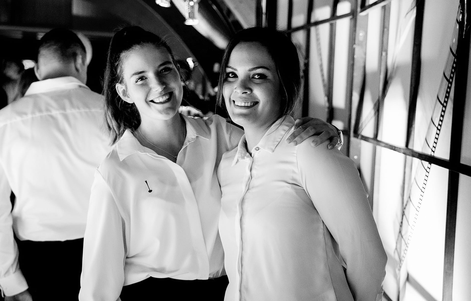 Zwei Frauen in hellen Blusen stehen nebeneinander und lächeln in die Kamera; sie befinden sich in einem gut beleuchteten Innenraum, im Hintergrund sind weitere Personen zu sehen.