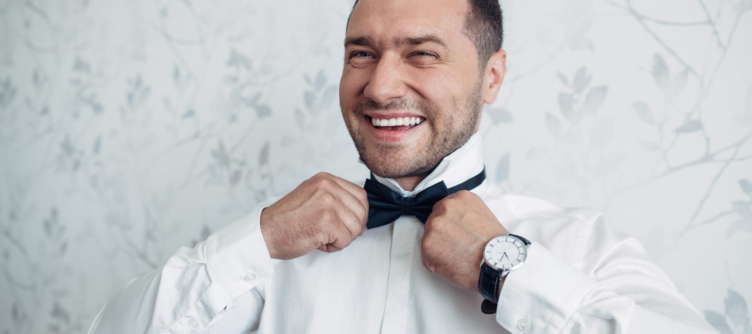 Ein lächelnder Mann in einem weißen Hemd, der sich eine schwarze Fliege richtet. Er trägt eine Armbanduhr mit dunklem Lederarmband und blickt freudig zur Seite. Der Hintergrund ist hell mit einem dezenten floralen Muster.