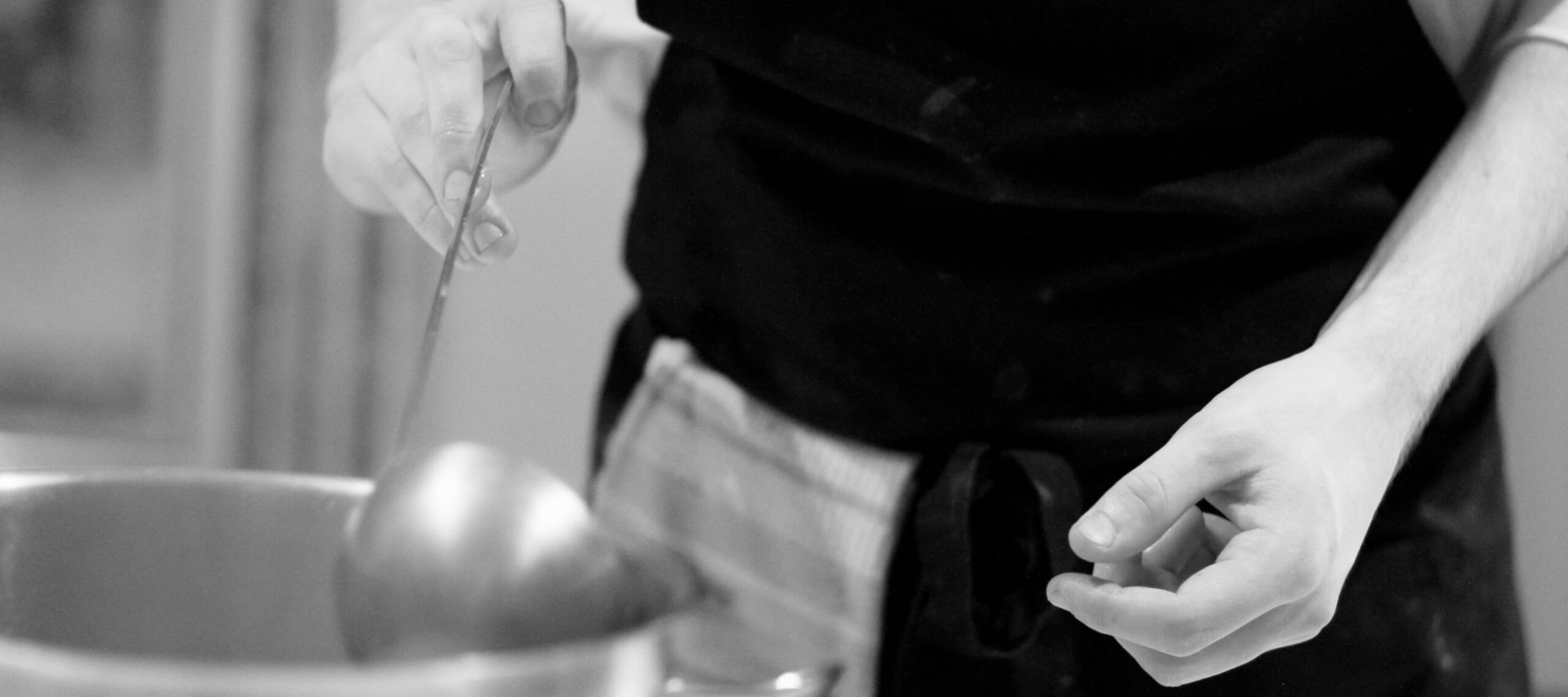 Eine Person, die eine Schürze trägt, hält eine Kelle über einen Topf und bereitet in einer Küche Essen zu. Das Bild ist in Schwarz-Weiß.